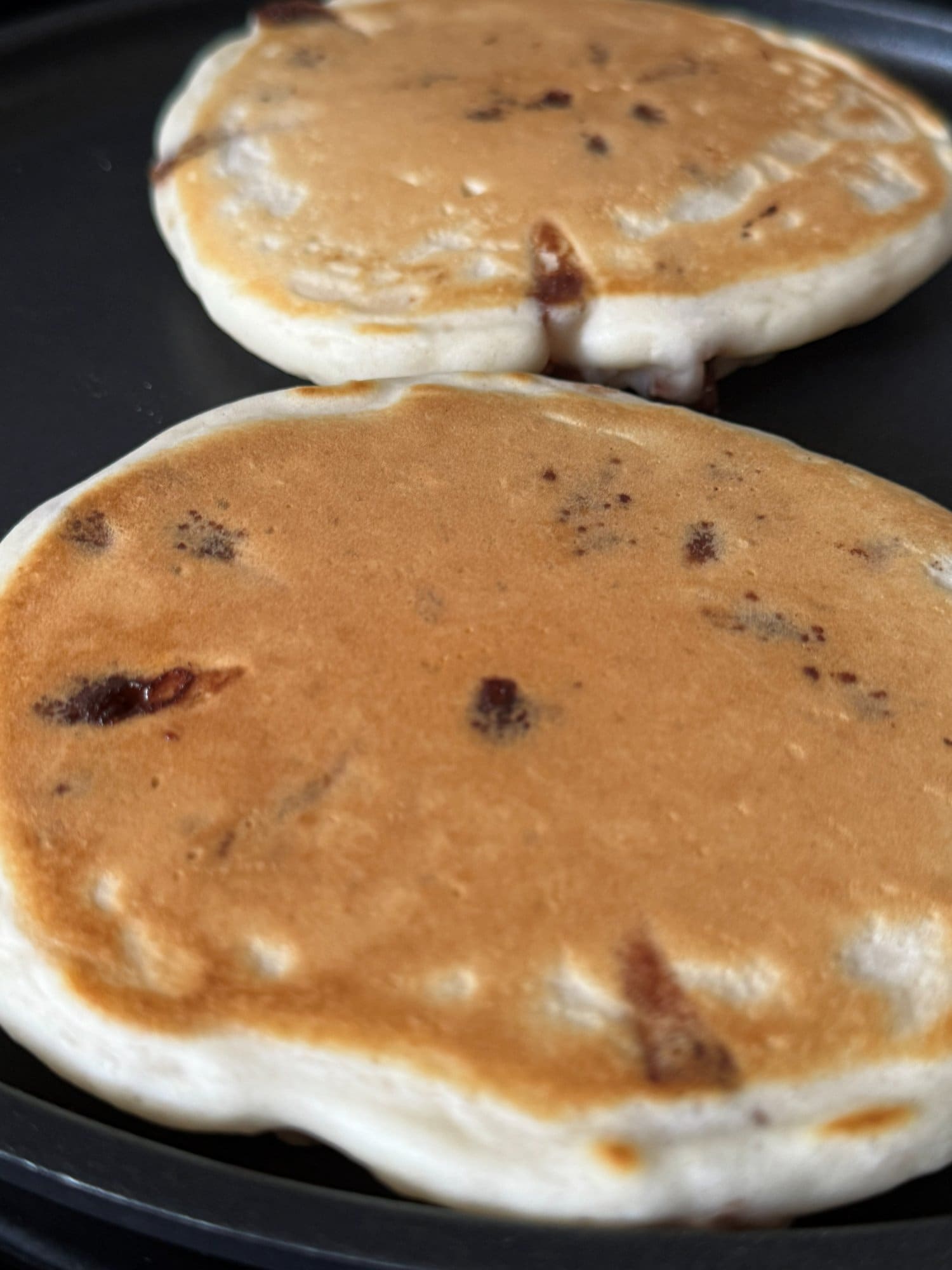 Two chocolate chip pancakes cooking on a griddle, the undersides golden brown